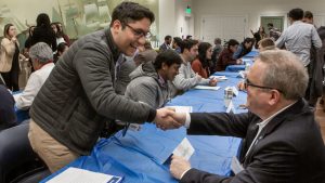 Two people shaking hands over a table and with people in the background.