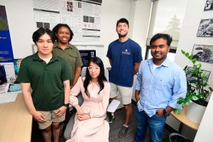 Jean Fan sits in her desk chair, surround by 4 members of her grad students. On the wall are various posters of cell images and formulas.